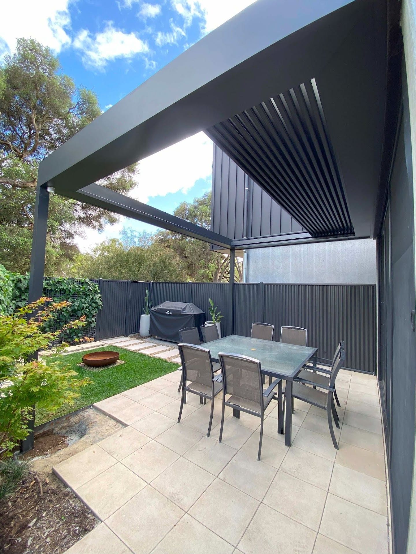 Modern outdoor deck with an adjustable black louvre roof, tiled patio, outdoor dining furniture and garden views.
