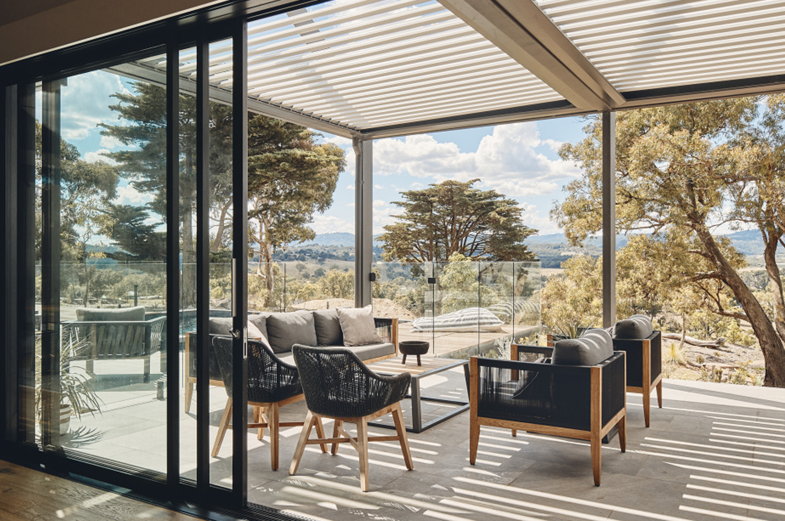 A luxurious patio with outdoor furniture under a louvered pergola, overlooking a scenic landscape.