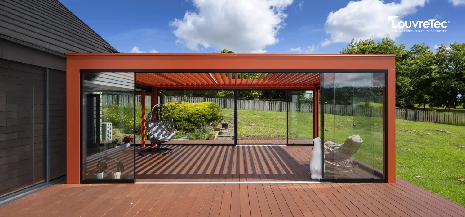 Outdoor louvre roof with glass walls on a wooden deck, overlooking a fenced garden.