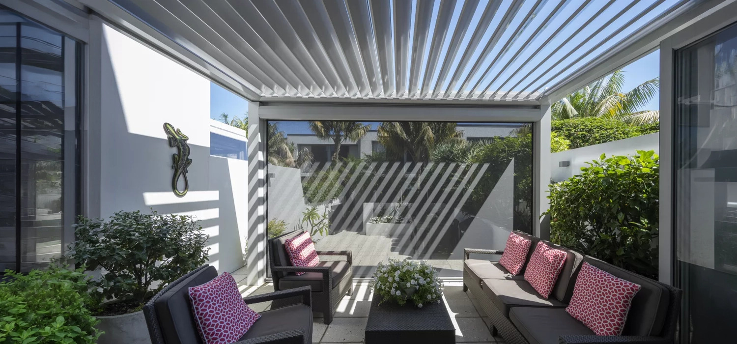 Outdoor louvre roof over a patio seating area, casting striped shadows across sofas and plants.