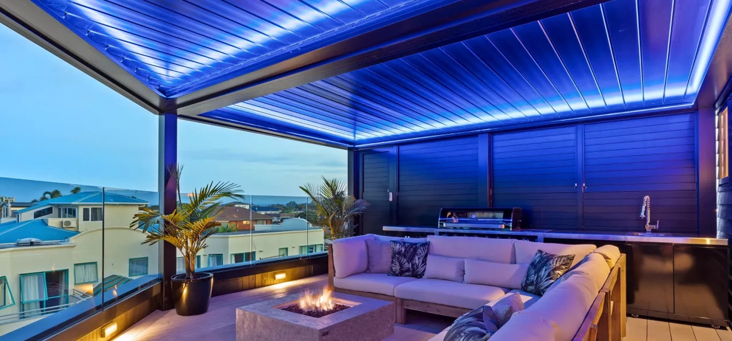 Outdoor louvre roof with blue lighting over a terrace sofa and fire pit, overlooking rooftops.