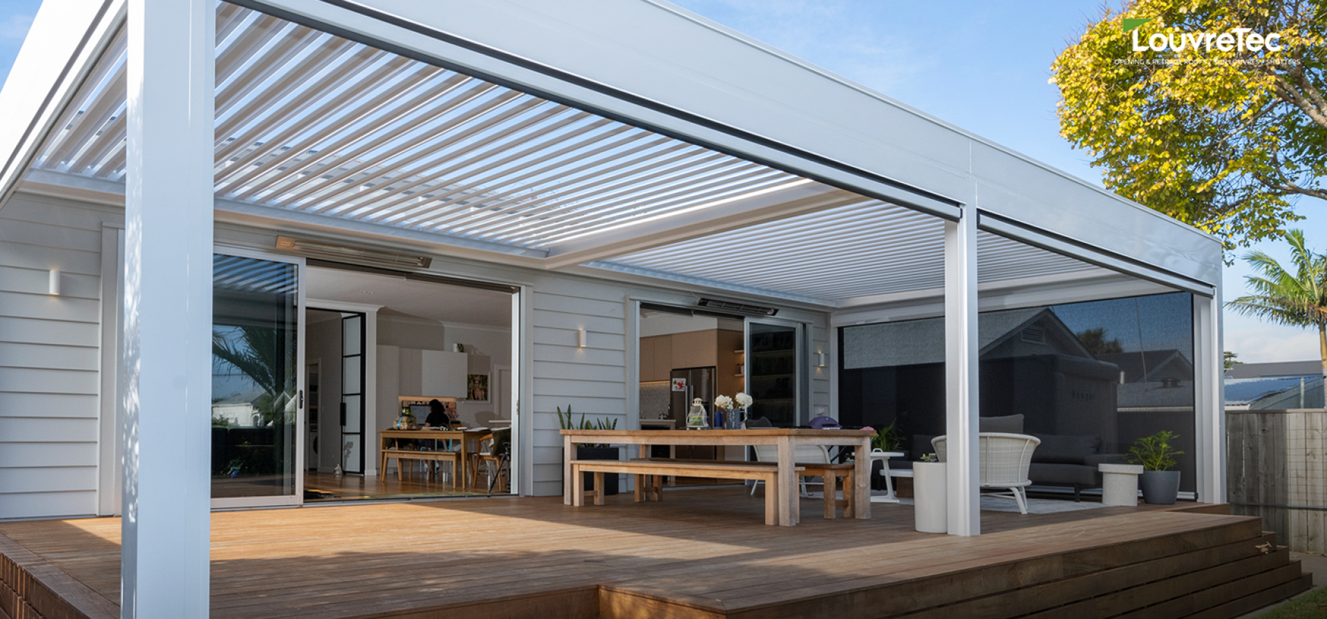 Opening roof system with adjustable louvered blades covering a timber deck and outdoor dining area attached to a modern home