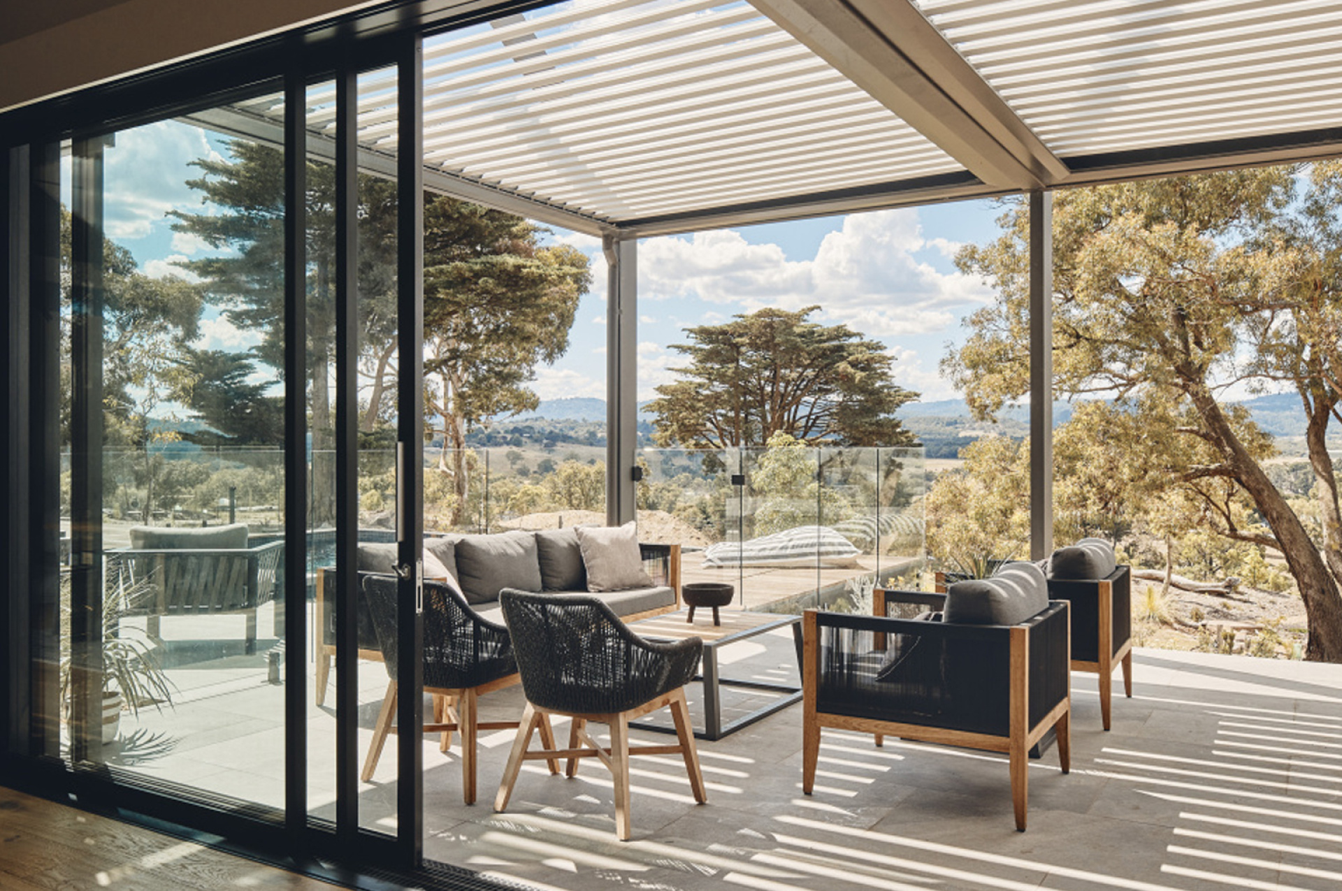 A luxurious patio with outdoor furniture under a louvered pergola, overlooking a scenic landscape.