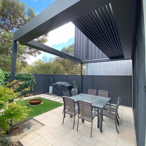 Modern outdoor deck with an adjustable black louvre roof, tiled patio, outdoor dining furniture and garden views.
