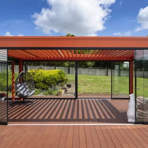 Outdoor louvre roof with glass walls on a wooden deck, overlooking a fenced garden.