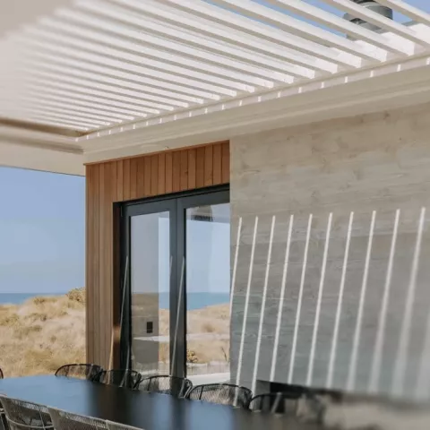 Outdoor dining area with table and chairs in front of a large window, shaded by a Louvretec louvre roof.