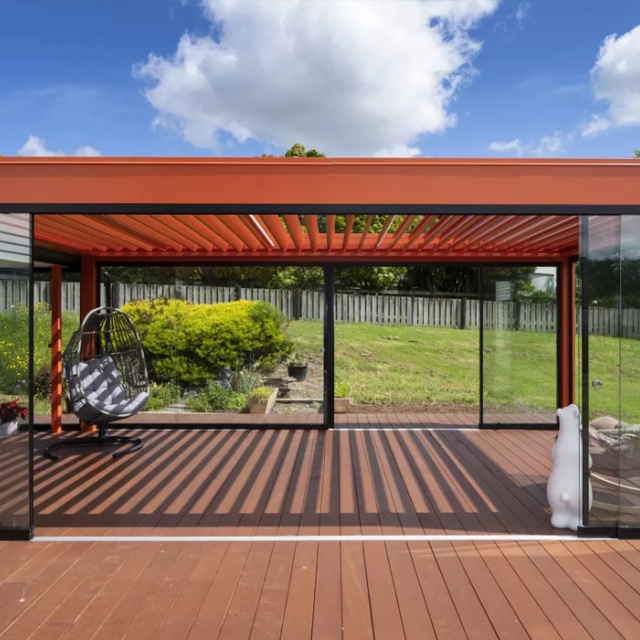 Outdoor louvre roof with glass walls on a wooden deck, overlooking a fenced garden.