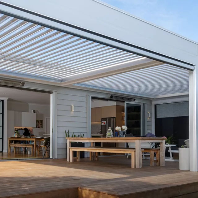 Opening roof system with adjustable louvered blades covering a timber deck and outdoor dining area attached to a modern home